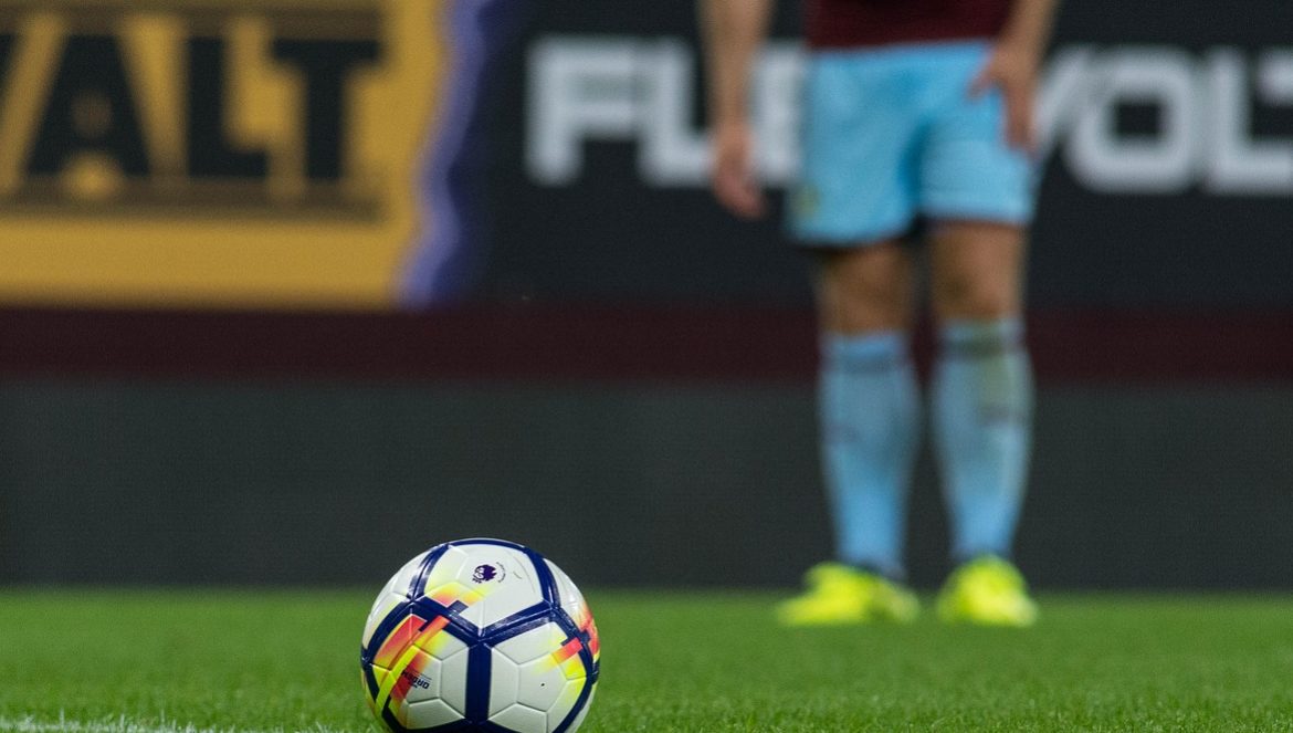 A soccer ball on grass with a player standing in the background.